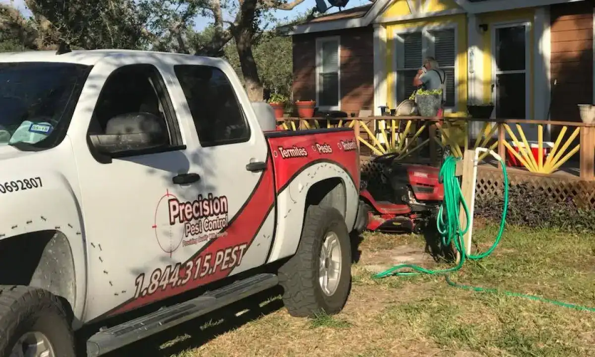 A pest control truck from Precision Pest Control parked near a yellow and brown house, with a worker preparing equipment on the lawn.