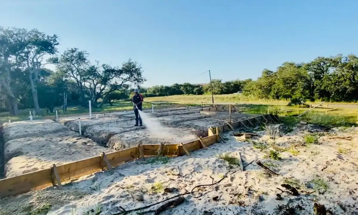 A worker spraying termite treatment on the sandy foundation of a construction site surrounded by trees and open space.