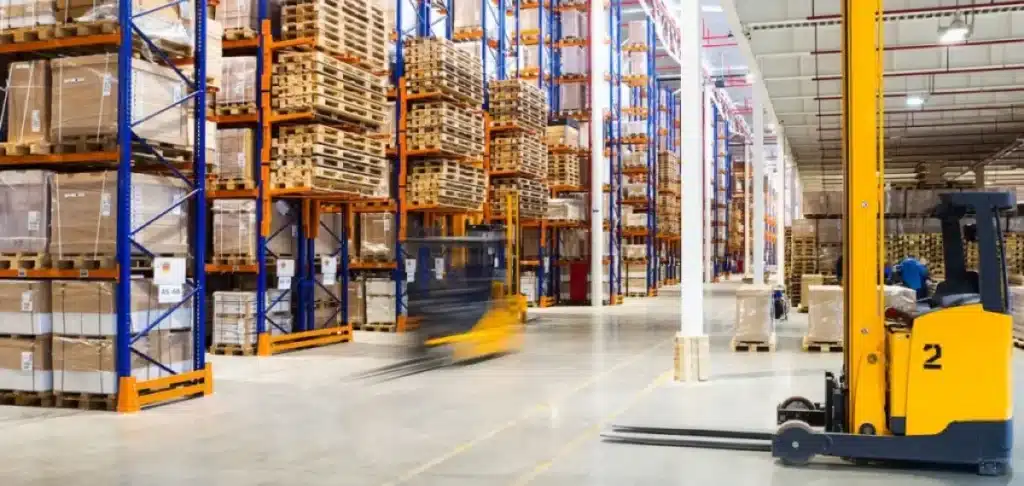 A large warehouse with tall blue and orange racks filled with wooden pallets and boxes, a forklift in motion, and bright lighting.