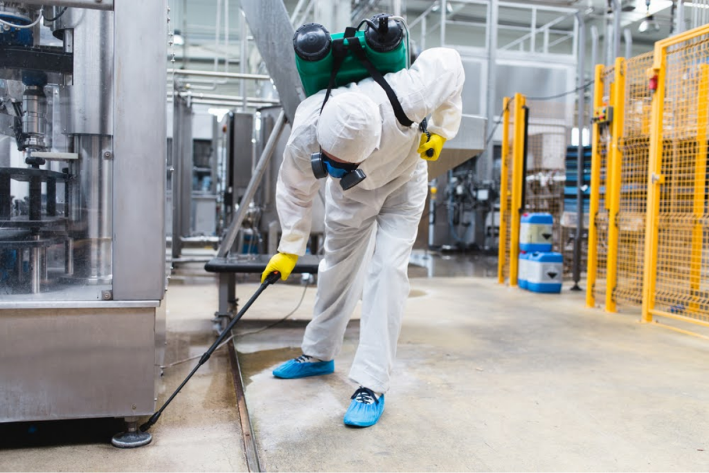 A person in full protective gear and a respirator cleaning a factory floor with specialized equipment, surrounded by industrial machines.