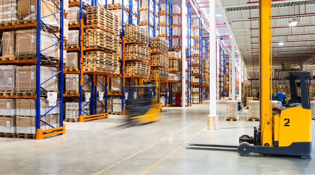 A large warehouse with tall blue and orange racks filled with wooden pallets and boxes, a forklift in motion, and bright lighting.