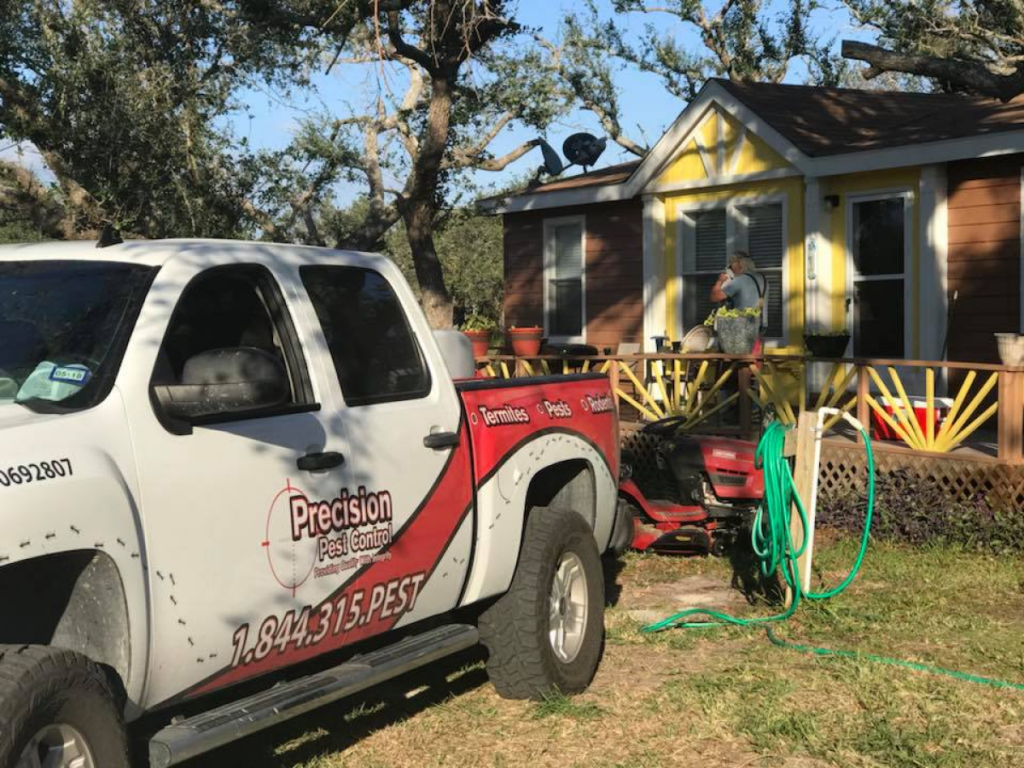 A pest control truck from Precision Pest Control parked near a yellow and brown house, with a worker preparing equipment on the lawn.