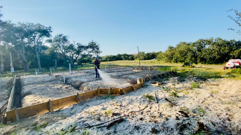 A worker spraying termite treatment on the sandy foundation of a construction site surrounded by trees and open space.