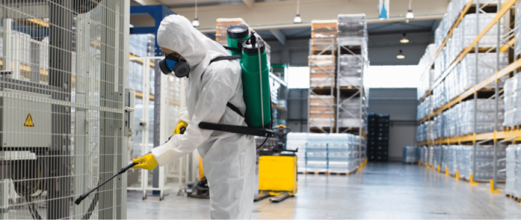 A worker in protective clothing disinfecting a warehouse area with shelves and stacked pallets using a spray applicator.